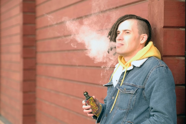 Foto retrato de joven guapo con rastas vapeando de pie apoyado en la pared en la calle de la ciudad adulto chico fumando ecigarette desahogándose