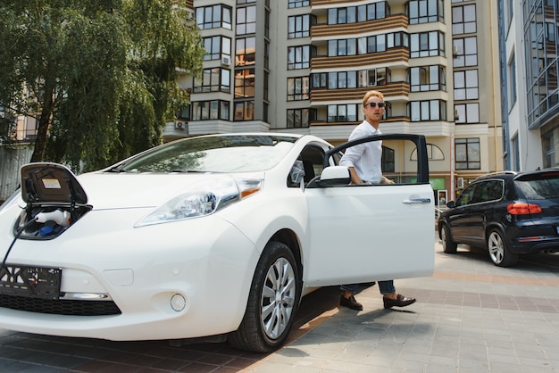 Retrato de joven guapo en ropa casual, de pie en la estación de carga. Concepto de coche eléctrico ecológico