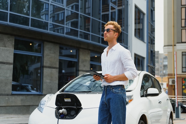 Retrato de joven guapo en ropa casual, de pie en la estación de carga. Concepto de coche eléctrico ecológico