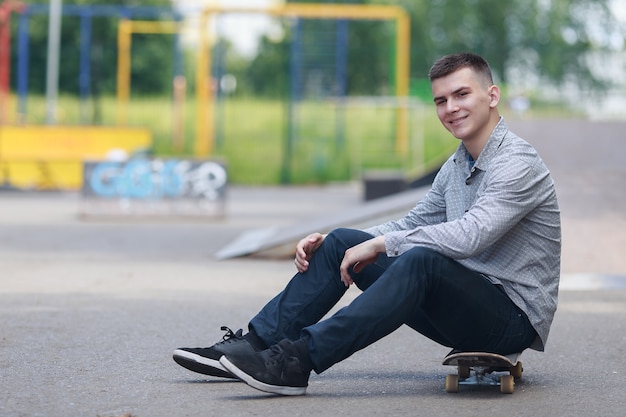 Retrato de joven guapo sonriente con ropa elegante sentado en la pizarra mientras se relaja en el parque de verano en medio de árboles verdes borrosos y barras horizontales deportivas