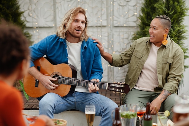 Retrato de joven guapo tocando la guitarra y cantando mientras disfruta de una fiesta al aire libre con amigos, espacio de copia