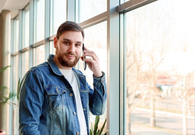 Retrato joven haciendo llamada. Hombre barbudo caucásico en la oficina de la ciudad moderna tiene conversación móvil