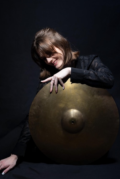 Retrato de una joven y hermosa baterista sonriente sosteniendo un gran platillo de tambor sobre fondo negro