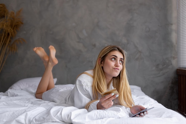 Retrato de una joven y hermosa chica caucásica acostada en la cama en casa comprando en línea