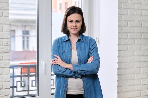 Retrato de una joven hermosa y confiada mujer sonriente con camisa de denim con los brazos cruzados, mujer mirando la cámara en casa