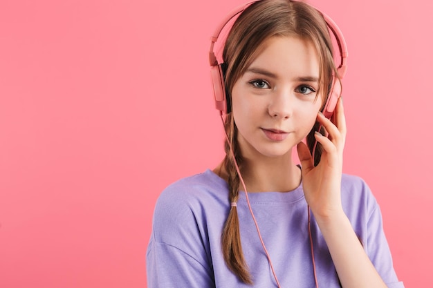 Retrato de una joven hermosa con dos trenzas en una camiseta lila escuchando música en los auriculares mirando soñadoramente a la cámara sobre un fondo rosa aislado