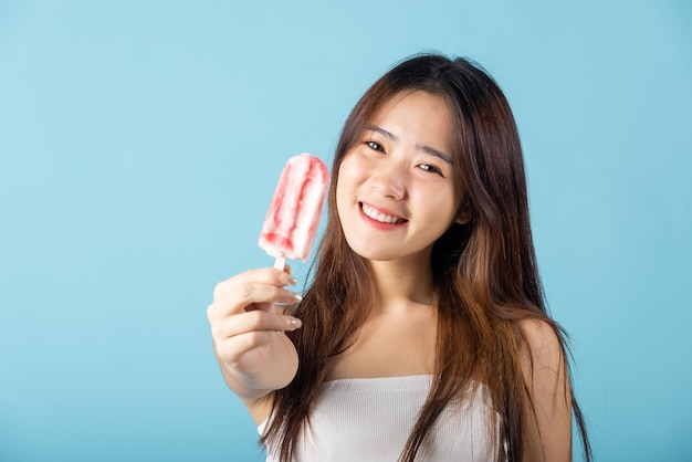 Retrato de una joven y hermosa mujer asiática feliz que sostiene un delicioso helado de madera con sabor a fruta mixta, una toma de estudio aislada en el fondo azul, un dulce y sabroso postre helado en verano