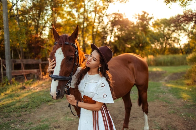 Retrato de una joven hermosa mujer con cabello rizado que sostiene un caballo marrón Caminando con los animales en la granja Montar a caballo jinete estilo boho