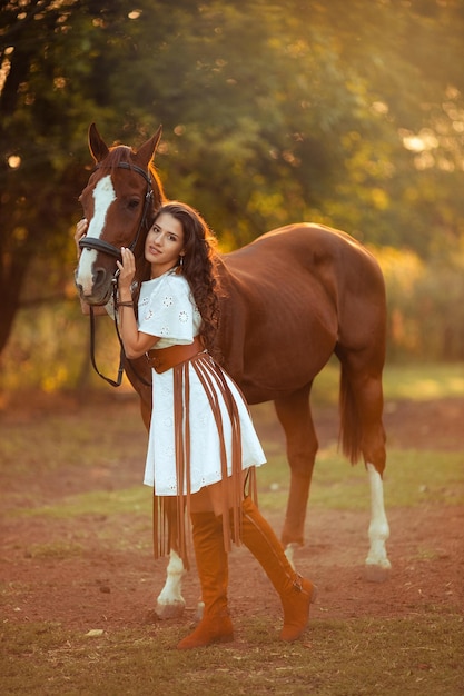 Foto retrato de una joven hermosa mujer con cabello rizado que sostiene un caballo marrón caminando con los animales en la granja montar a caballo jinete estilo boho