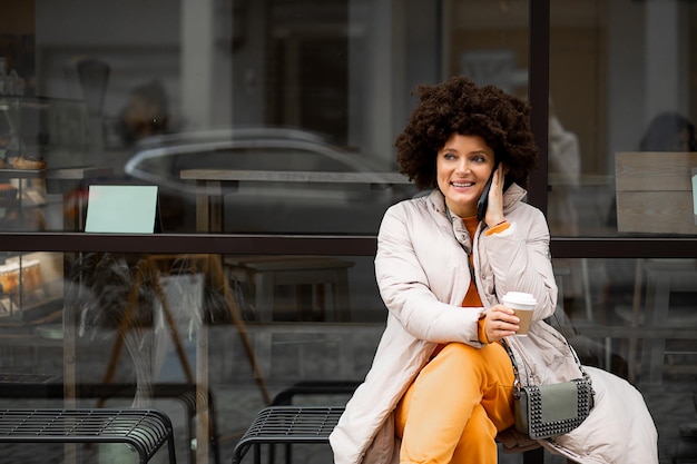 Retrato de joven, hermosa mujer caucásica de 40 años, ropa casual deportiva, sentado en un banco en la calle, hablando por teléfono inteligente, peinado afro. Disfrutando de la vida en otoño. Vida en la ciudad