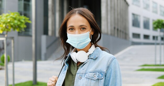 Retrato de joven hermosa mujer caucásica en máscara médica mirando a la cámara al aire libre y riendo. Estudiante alegre atractiva en auriculares y con la espalda en la calle en cuarentena. Pandemia.