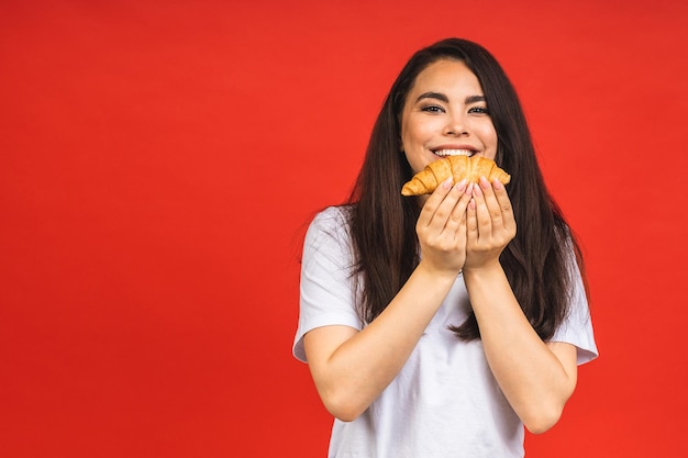 Retrato de joven hermosa mujer hambrienta comiendo croissant Retrato aislado de mujer con comida rápida sobre fondo rojo Concepto de desayuno dietético
