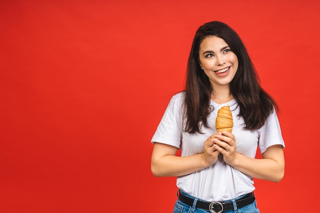 Retrato de joven hermosa mujer hambrienta comiendo croissant Retrato aislado de mujer con comida rápida sobre fondo rojo Concepto de desayuno dietético