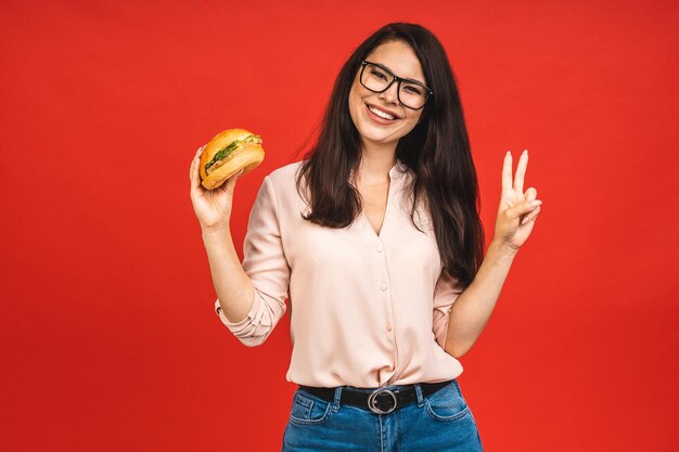 Retrato de joven hermosa mujer hambrienta comiendo hamburguesa Retrato aislado de estudiante con comida rápida sobre fondo rojo Concepto de dieta