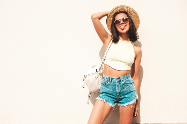 Retrato de joven hermosa mujer hipster sonriente en pantalones cortos de jeans de verano de moda y sombrero Modelo despreocupado sexy posando en la calle cerca de la pared blanca Modelo positivo al aire libre con maletínMuestra lengua
