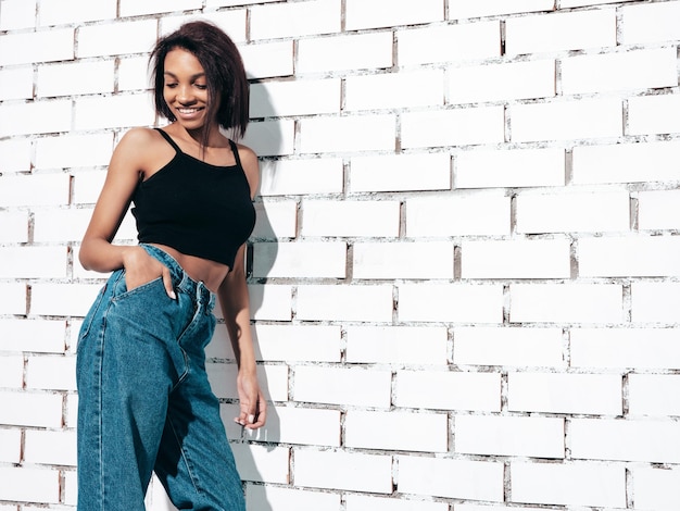 Retrato de joven hermosa mujer negra Modelo sonriente vestida con jeans de verano y ropa superior negra Sexy mujer despreocupada posando junto a la pared de ladrillo blanco en el estudio Bronceada y alegre