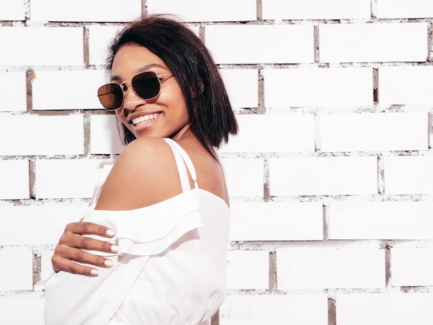 Retrato de joven hermosa mujer negra Modelo sonriente vestida con ropa de jeans de verano Sexy mujer despreocupada posando junto a la pared de ladrillo blanco en el estudio Bronceada y alegre En gafas de sol