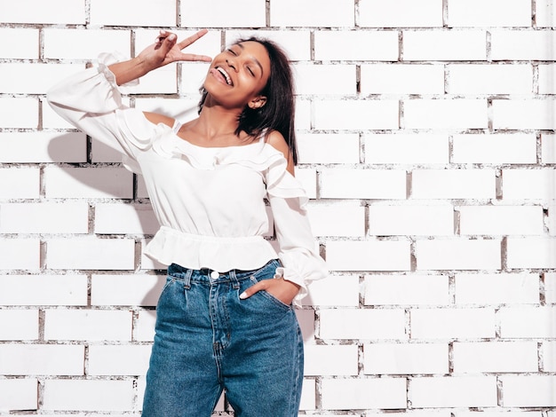 Retrato de joven hermosa mujer negra Modelo sonriente vestida con ropa de jeans de verano Sexy mujer despreocupada posando junto a la pared de ladrillo blanco en el estudio Bronceada y alegre Muestra el signo de la paz y la lengua