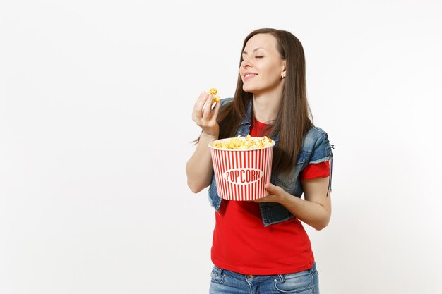 Retrato de joven hermosa mujer relajante con los ojos cerrados en ropa casual viendo una película, sosteniendo un cubo de palomitas de maíz, comiendo, disfrutando aislado sobre fondo blanco. Emociones en el concepto de cine.