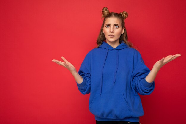 Retrato de joven hermosa mujer rubia atractiva con dos cuernos con emociones sinceras vistiendo elegante sudadera con capucha azul brillante aislada en la pared roja con espacio de copia y tener dudas.