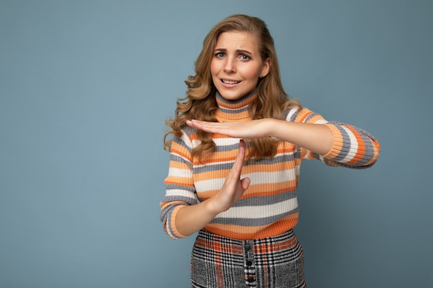 Retrato de joven hermosa mujer rubia rizada con emociones sinceras vistiendo elegante suéter a rayas aislado sobre fondo azul con espacio vacío y mostrando gesto de tiempo de espera