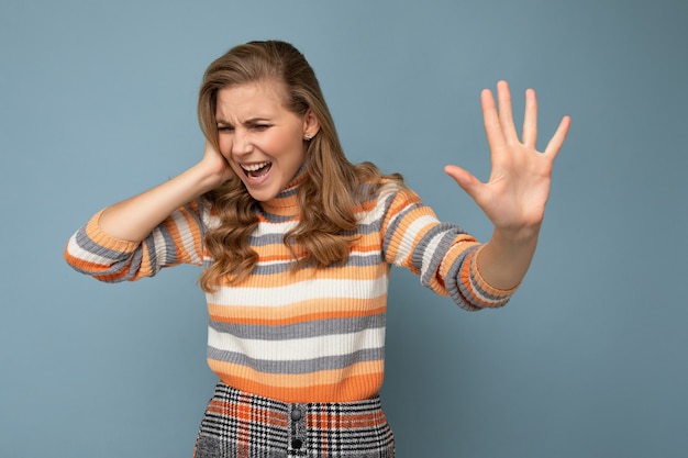 Retrato de joven hermosa mujer rubia rizada con emociones sinceras vistiendo elegante suéter a rayas aislado sobre fondo azul con espacio vacío y mostrando la palma de la mano con expresión repugnante