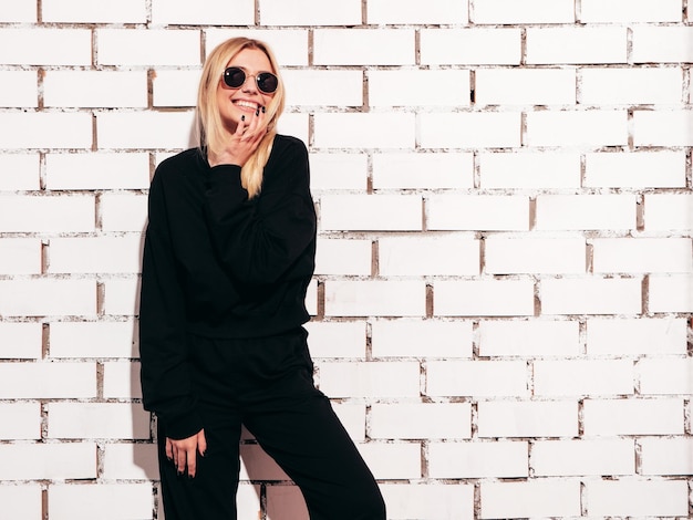 Retrato de joven hermosa mujer rubia sonriente en ropa de gran tamaño negra de verano de moda Mujer despreocupada sexy posando junto a la pared de ladrillo blanco en el estudio Modelo positivo divirtiéndose en el interior En gafas de sol