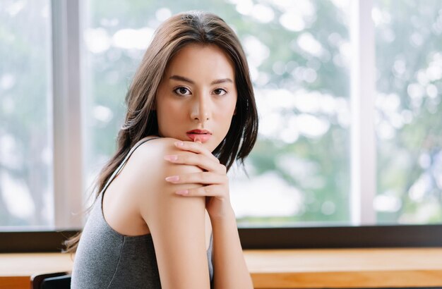 Retrato de una joven hermosa mujer sentada en una silla con la mano en el hombro y mirando la cámara con fondo verde a través de la ventana de la sala de estar con espacio para copiar