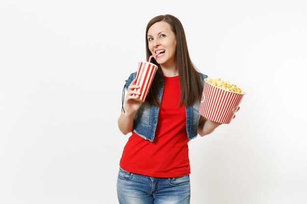 Retrato de joven hermosa mujer sonriente en ropa casual viendo una película, sosteniendo un cubo de palomitas de maíz, bebiendo de un vaso de plástico de refresco o cola aislado sobre fondo blanco. Emociones en el cine.