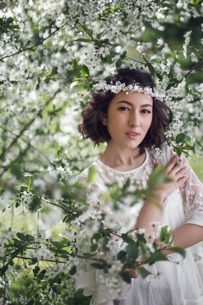 Retrato de una joven hermosa mujer vestida de blanco parada al lado de un cerezo floreciente en primavera una corona en la cabeza