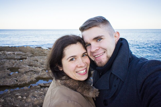 Retrato de joven hermosa pareja tomando foto selfie en un teléfono inteligente con mar y cielo nublado oscuro en el fondo. Temporada fría y concepto de viaje.