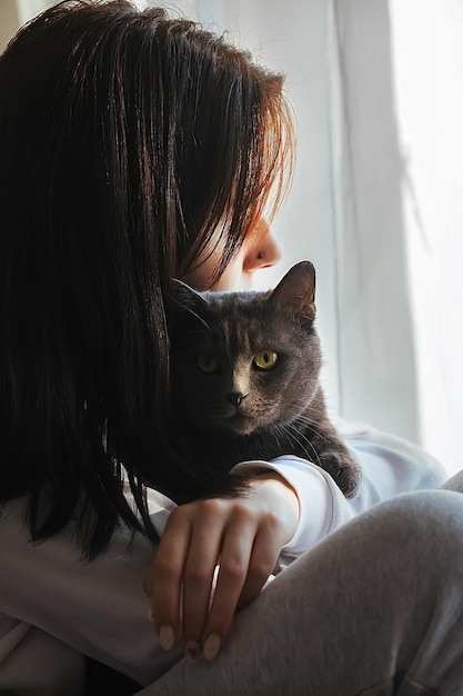 Retrato de una joven hermosa con su gato gris cerca de la ventana contraste de luz y sombra