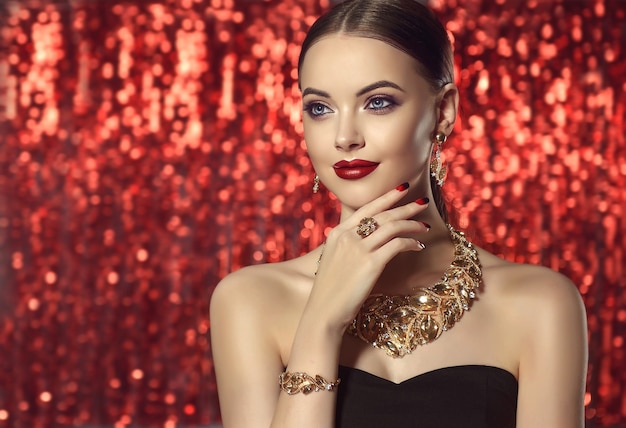 Retrato de joven hermosa vestida con un conjunto de joyas de collar, anillo, pulsera y aretes. La modelo de ojos azules está demostrando un atractivo maquillaje y manicura sobre el fondo rojo brillante.