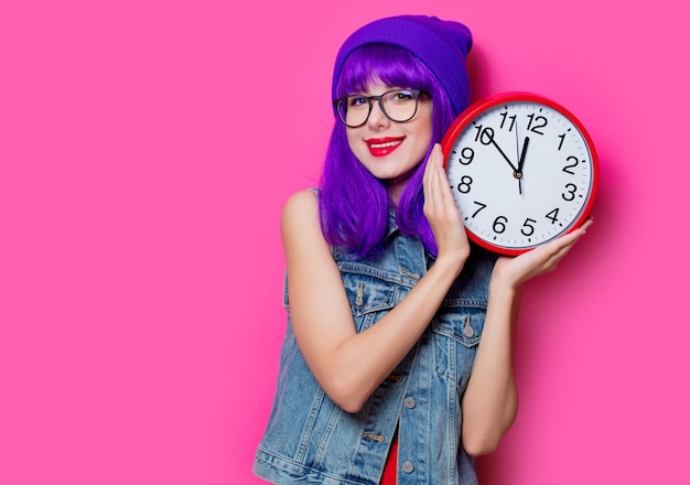 Retrato de una joven hipster de estilo con cabello morado y un gran reloj sobre fondo rosa