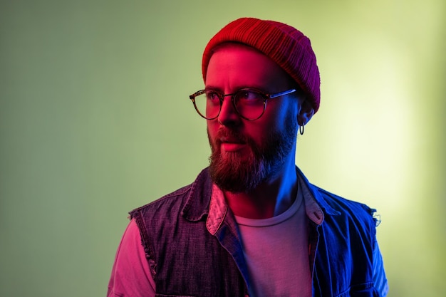 Retrato de un joven hipster de moda con anteojos posando y mirando hacia otro lado con una cara seria y pensativa, usando sombrero de beanie y chaleco de mezclilla. Disparo de estudio interior aislado sobre fondo de luz de neón colorido.