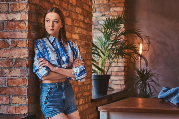 Retrato de una joven hipster vestida con una camisa y pantalones vaqueros cruzados de brazos mientras se apoyaba en una mesa en una habitación con interior de loft.