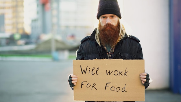 Retrato de un joven sin hogar con cartón mirando a la cámara y quiere trabajar por comida
