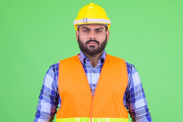 Retrato de joven hombre indio barbudo con sobrepeso, trabajador de la construcción contra chroma key con pared verde