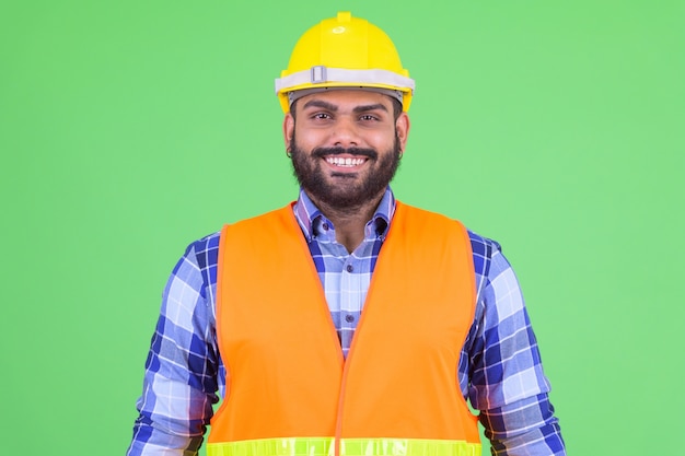 Retrato de joven hombre indio barbudo con sobrepeso, trabajador de la construcción contra chroma key con pared verde