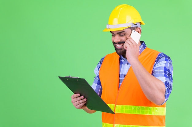 Foto retrato de joven hombre indio barbudo con sobrepeso, trabajador de la construcción contra chroma key con pared verde