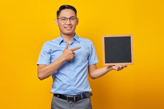 Retrato de un joven hombre de negocios asiático sonriente con anteojos señalando la pizarra con el dedo y de pie confiado mirando la cámara aislada sobre el fondo amarillo