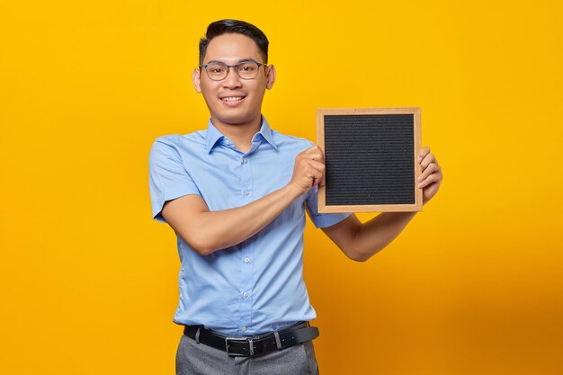 Retrato de un joven hombre de negocios asiático sonriente con anteojos sosteniendo pizarra y de pie confiado mirando la cámara aislada sobre el fondo amarillo