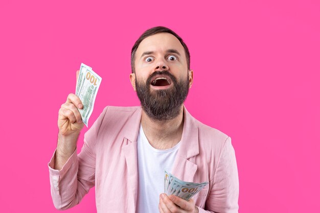 Retrato de un joven hombre de negocios contento con barba vestido con una chaqueta rosa que muestra billetes de dólar estadounidense contra un fondo de estudio rojo Gusto olor a dinero