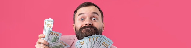 Retrato de un joven hombre de negocios contento con barba vestido con una chaqueta rosa que muestra billetes de dólar estadounidense contra un fondo de estudio rojo Gusto olor a dinero