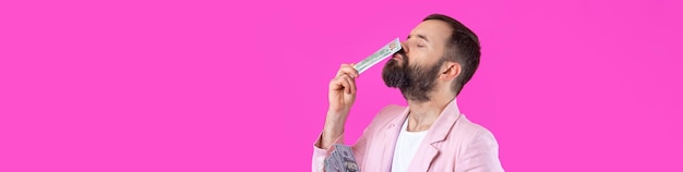 Retrato de un joven hombre de negocios contento con barba vestido con una chaqueta rosa que muestra billetes de dólar estadounidense contra un fondo de estudio rojo Gusto olor a dinero