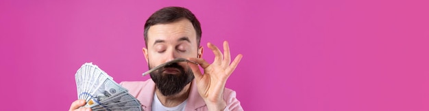 Retrato de un joven hombre de negocios contento con barba vestido con una chaqueta rosa que muestra billetes de dólar estadounidense contra un fondo de estudio rojo Gusto olor a dinero