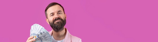 Retrato de un joven hombre de negocios contento con barba vestido con una chaqueta rosa que muestra billetes de dólar estadounidense contra un fondo de estudio rojo Gusto olor a dinero