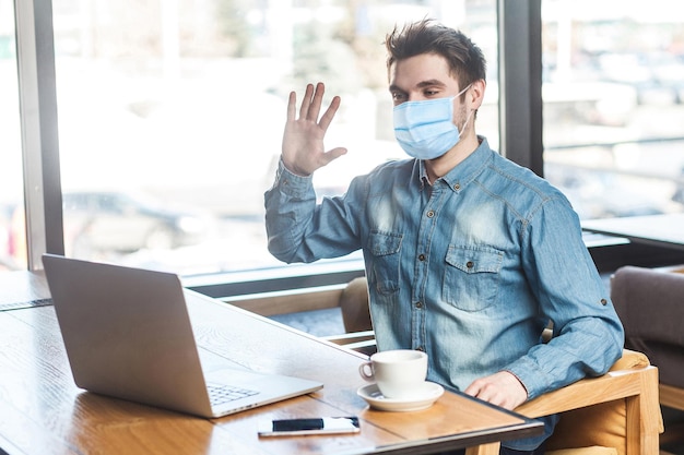 Foto retrato de un joven hombre de negocios feliz y satisfecho con una máscara médica quirúrgica en pantalones azul sentado y saludando a un trabajador a través de una cámara web en una reunión en línea. concepto de trabajo en interiores y atención médica.