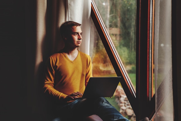 Retrato de un joven hombre de negocios con una laptop en la ventana de la sala de estar en una casa de campo. Hombre adicto al trabajo con ropa informal en casa trabajando de vacaciones. Inspiración creativa y puesta en marcha de empresas. copia espacio
