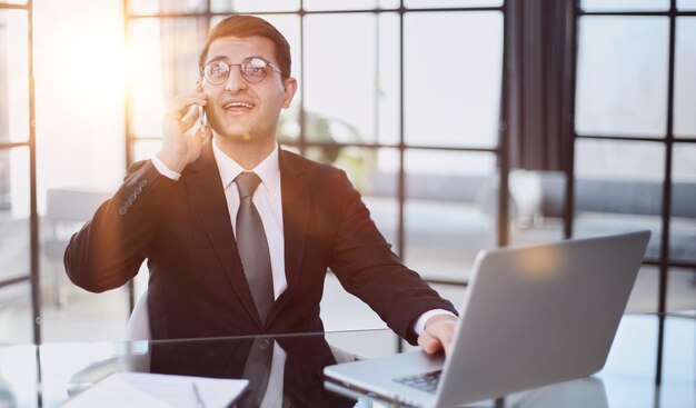 Retrato de un joven hombre de negocios serio en una oficina casual llamando por teléfono mientras trabaja con una computadora portátil